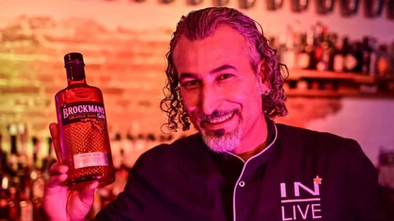 Barkeeper steht hinter einem beleuchteten Bartresen und hält eine Gin-Flasche in die Luft.