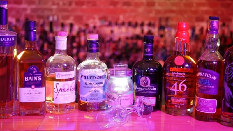 Whiskeyflaschen stehen aufgereiht in pinkem Licht auf einem Bartresen. Davor liegen zwei Gläser.