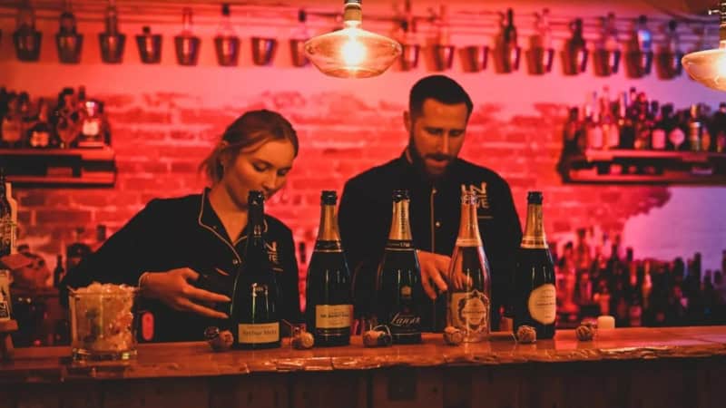 Ein Barkeeper und eine Barkeeperin stehen hinter einem Bartresen auf dem verschiedene Champagner-Flaschen präsentiert und für eine Verkostung vorbereitet werden.