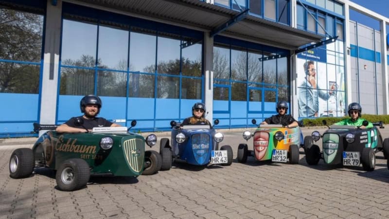 Eine Gruppe Hotrod Fahrer die in einem Halbkreis auf einem Parkplatz stehen.
