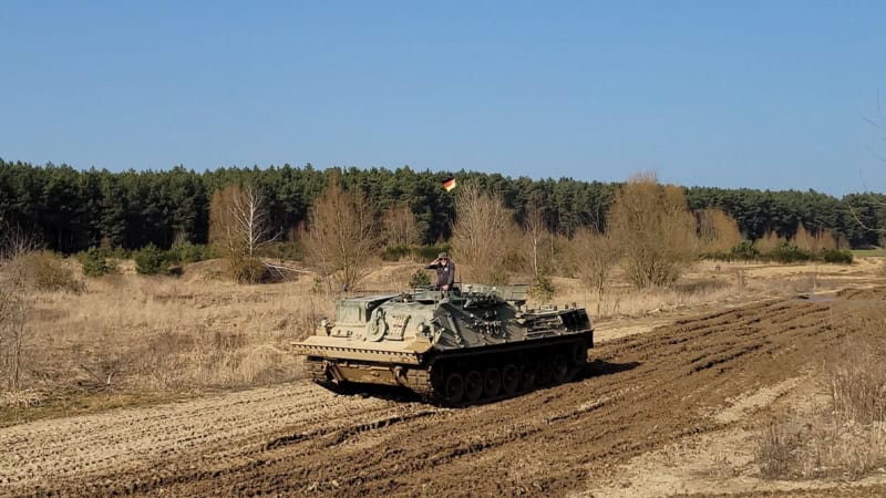 Ein Mann der mit einem Panzer über einen Matsch Weg fährt.